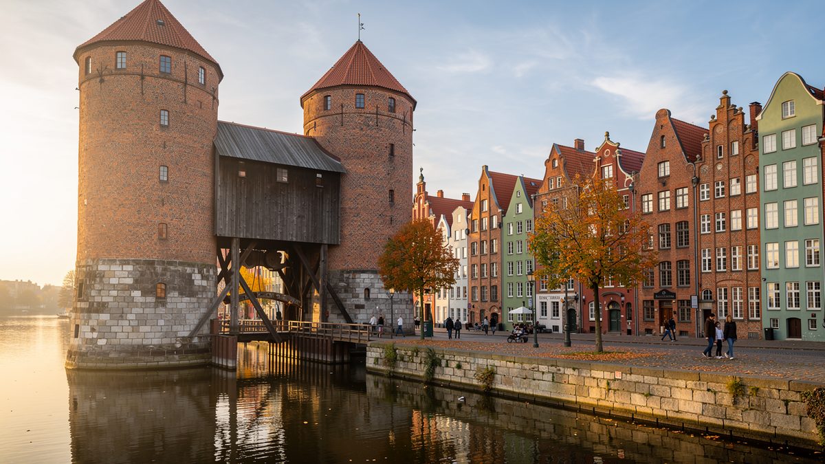 Two round brick towers with red roofs joined by a dark wooden crane housing over a river, next to a row of colourful gabled houses on a stone quay in autumn light (illustration)
