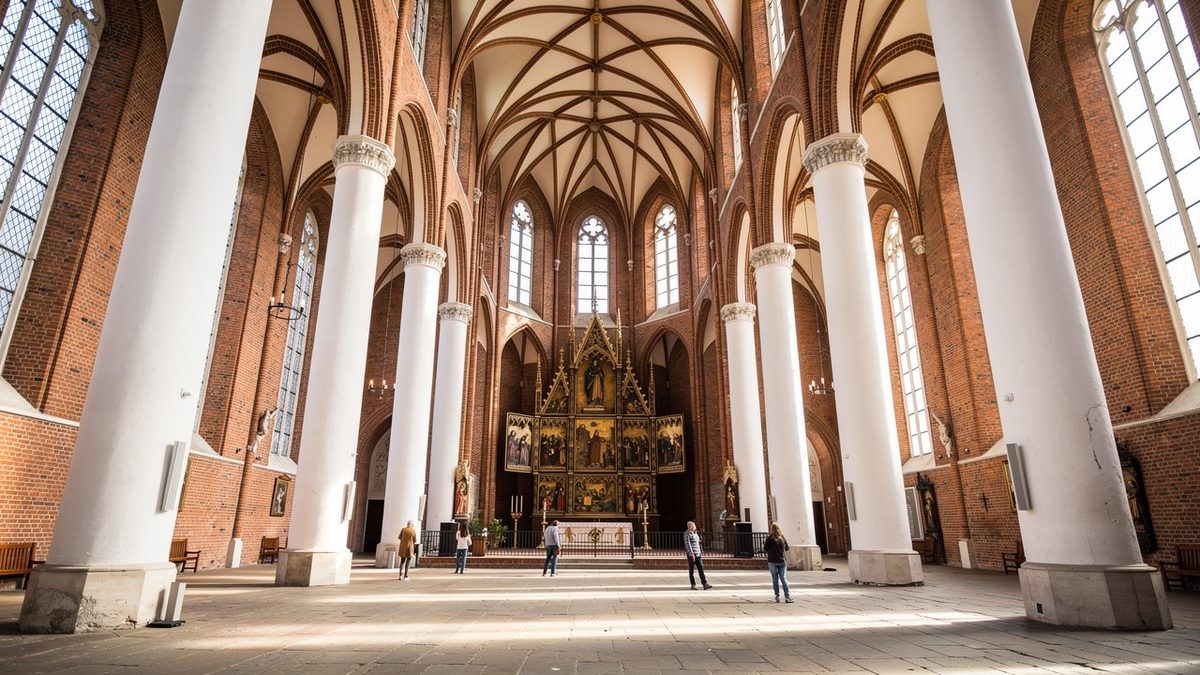 A tall Gothic brick hall church interior with white pillars, star vaulting and a carved winged altarpiece at the far end, with a few visitors on the stone floor (illustration)